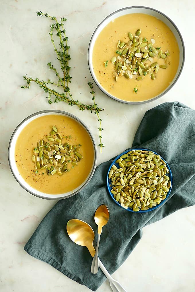 two bowls of butternut squash soup on a counter with pumpkin seeds, spoons, thyme, and a napkin