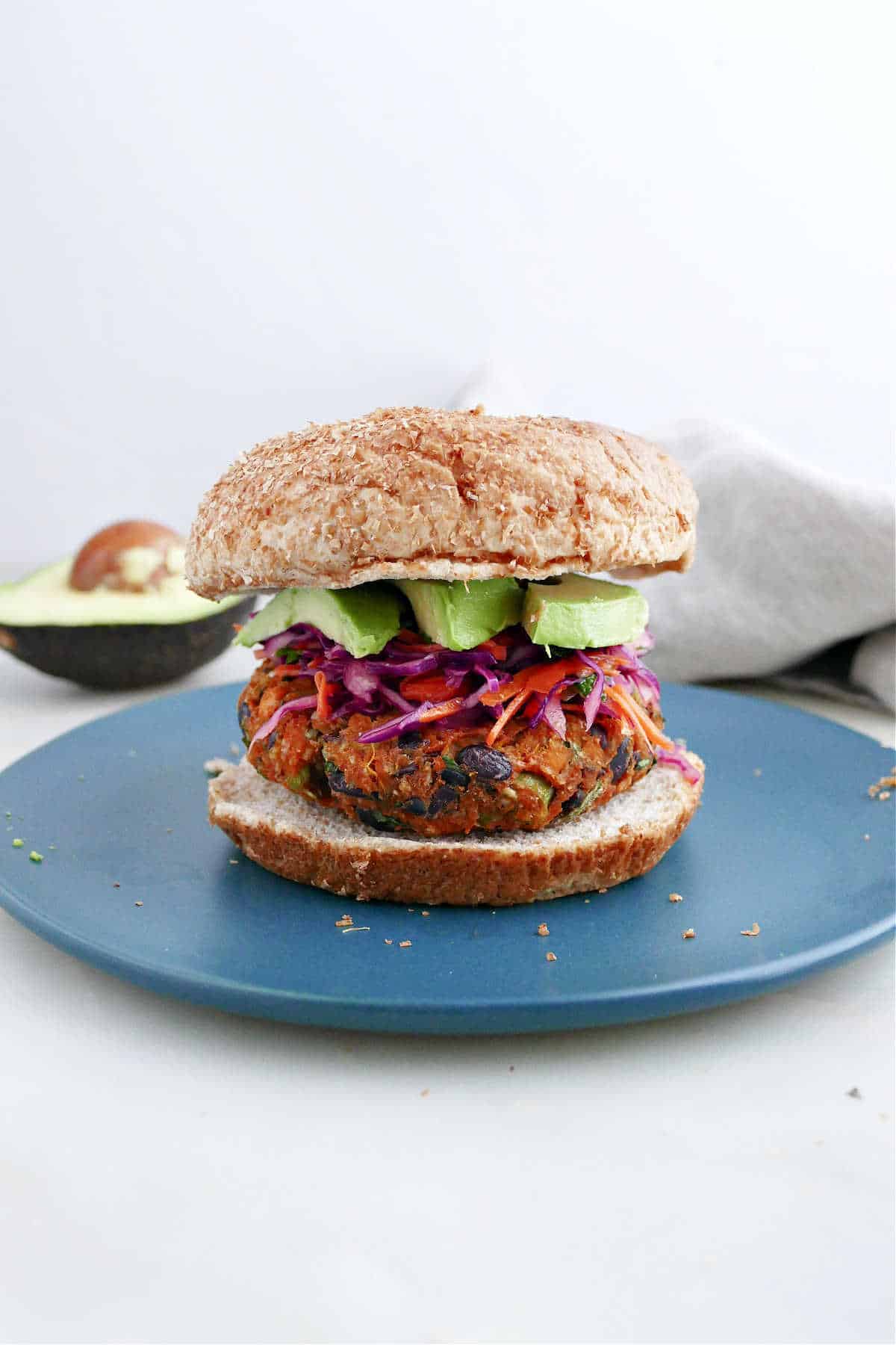 A sweet potato black bean burger with slaw and avocado slices on a blue plate.