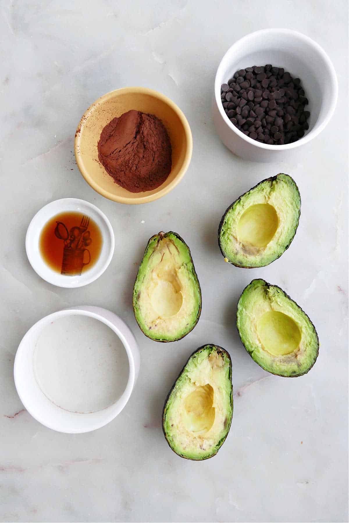 almond milk and maple syrup, cocoa powder, chocolate chips, vanilla, and avocados on a counter