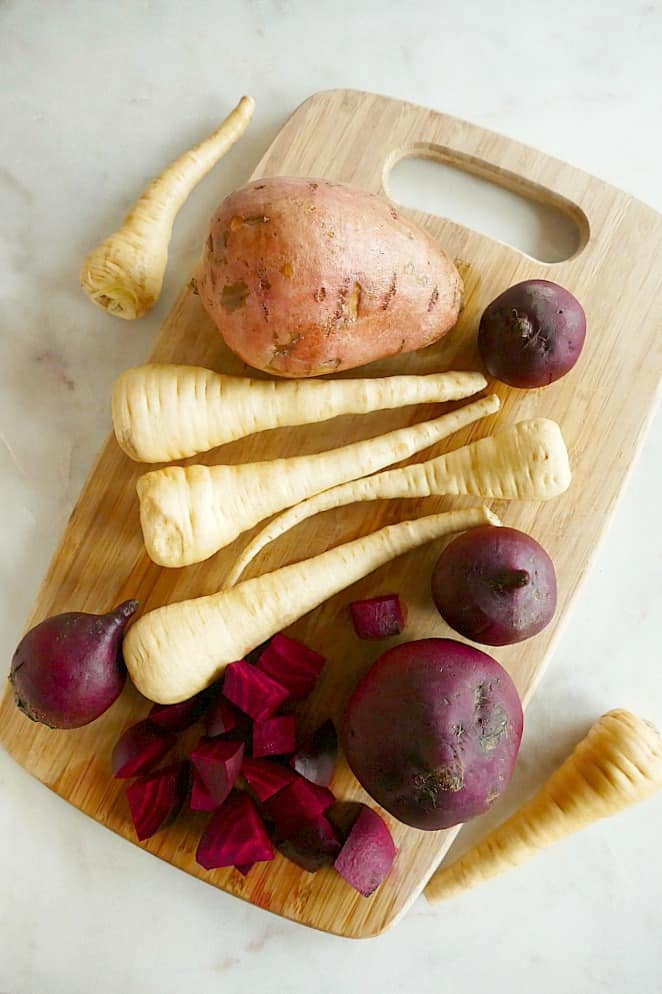 a bamboo cutting board with parsnips, beets, and a sweet potato on top