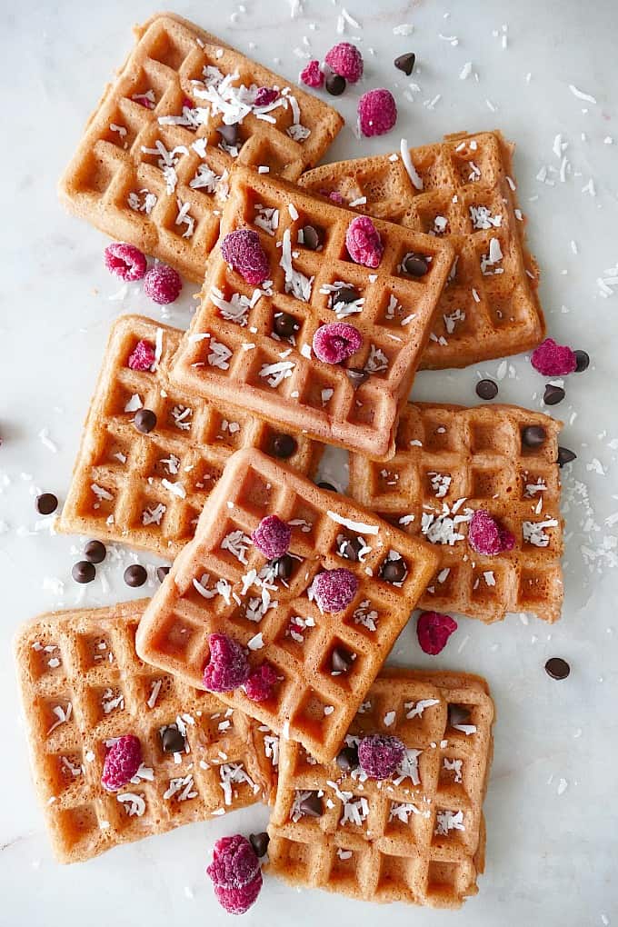 8 pink waffles on a white counter with shredded coconut, raspberries, and chocolate chips
