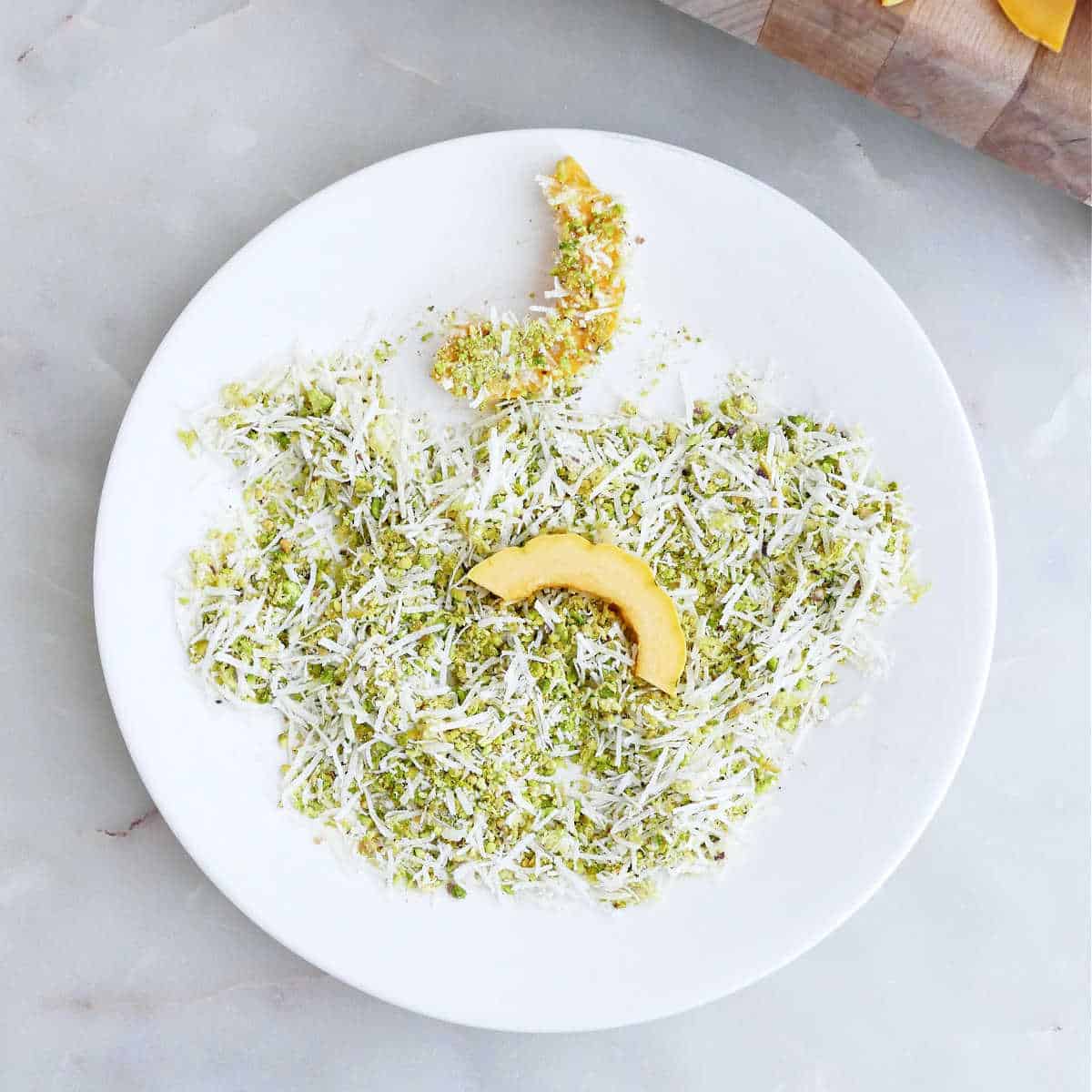 Delicata squash slices being pressed into ground pistachios and parmesan cheese on a plate.