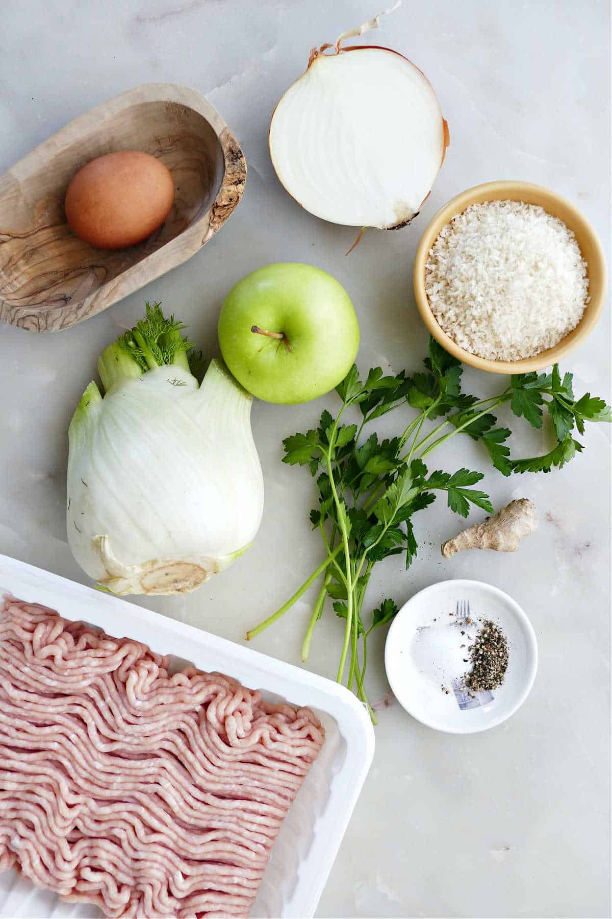 Ground chicken, fennel, apple, egg, onion, breadcrumbs, and seasonings on a counter.