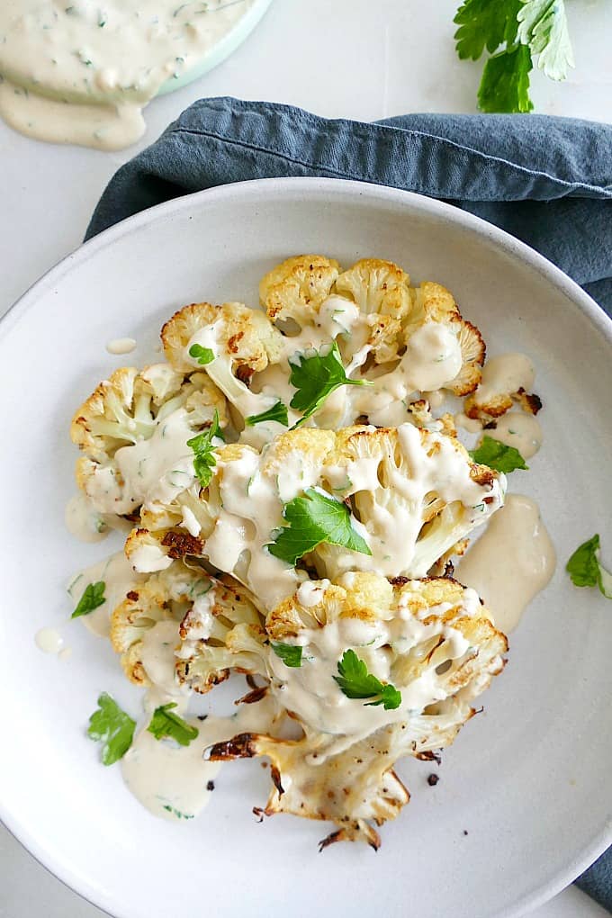 three cauliflower steaks with garlic tahini sauce on a white plate on a napkin