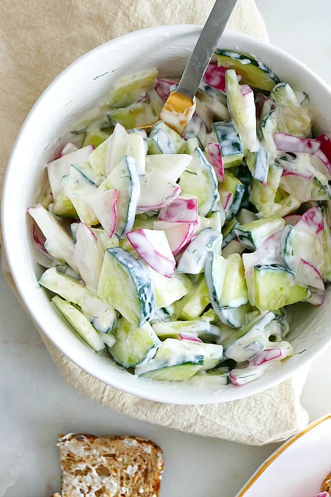 close up of sliced radishes and cucumbers coated in cream cheese in a white bowl