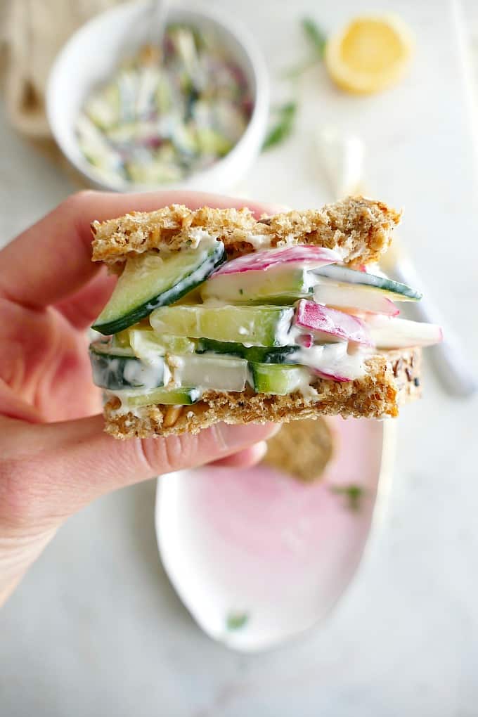hand holding one mini cucumber sandwich over a white counter with ingredients
