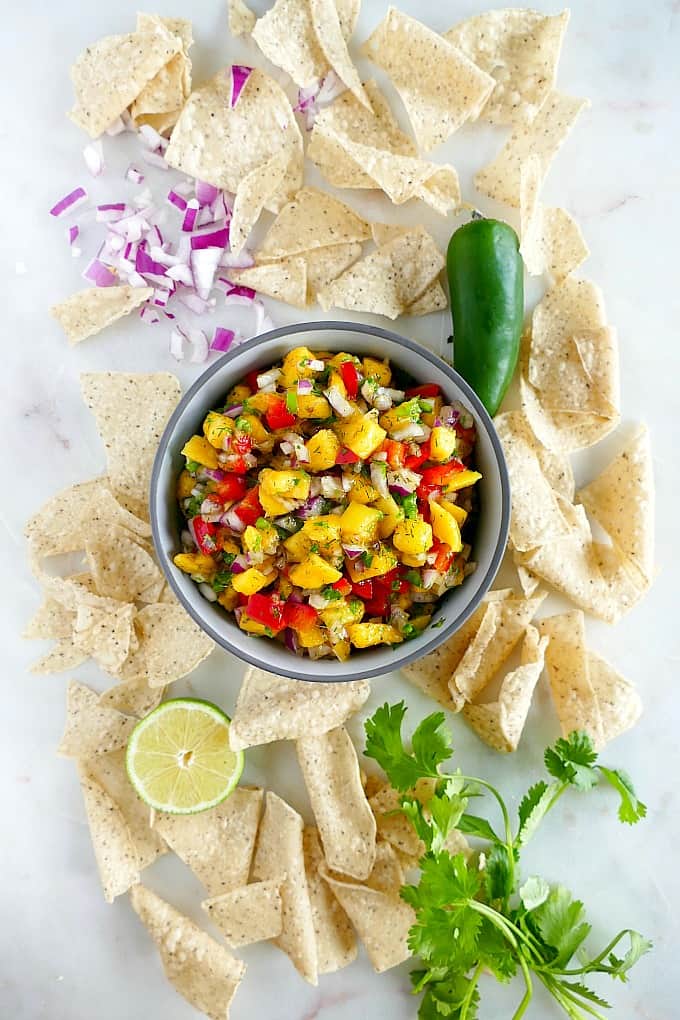 bowl of pepper mango salsa surrounded by ingredients and tortilla chips on a counter