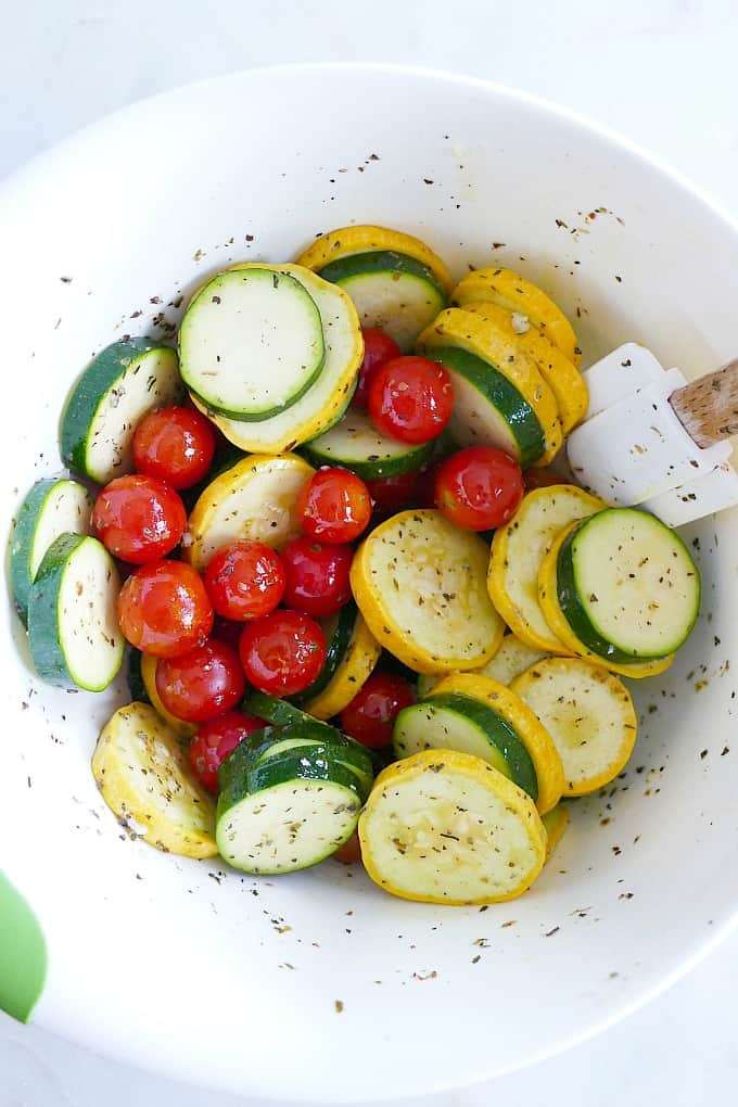 sliced summer squash and whole cherry tomatoes in a white mixing bowl with seasonings