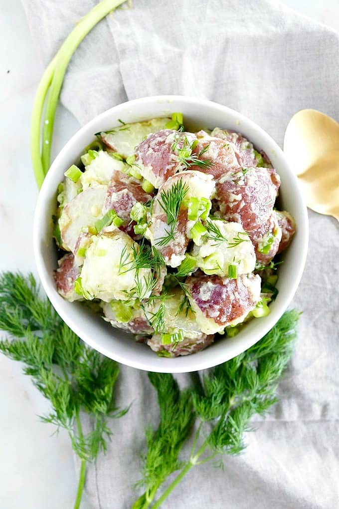 white bowl with potato salad on top of a gray napkin surrounded by fresh dill