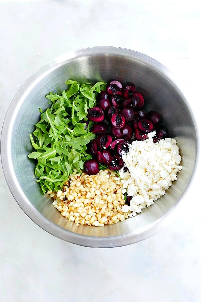 arugula, cherries, corn kernels, and feta cheese in mixing bowl on a counter