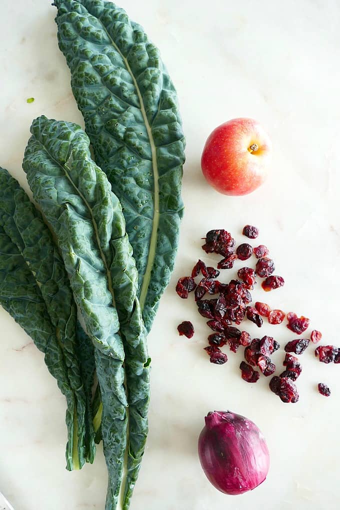 three long kale leaves, an apple, dried cranberries, and a red onion on a counter