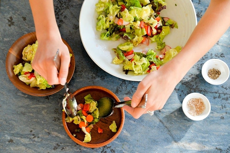 two hands tossing a salad in a brown bowl next to a larger bowl of salad