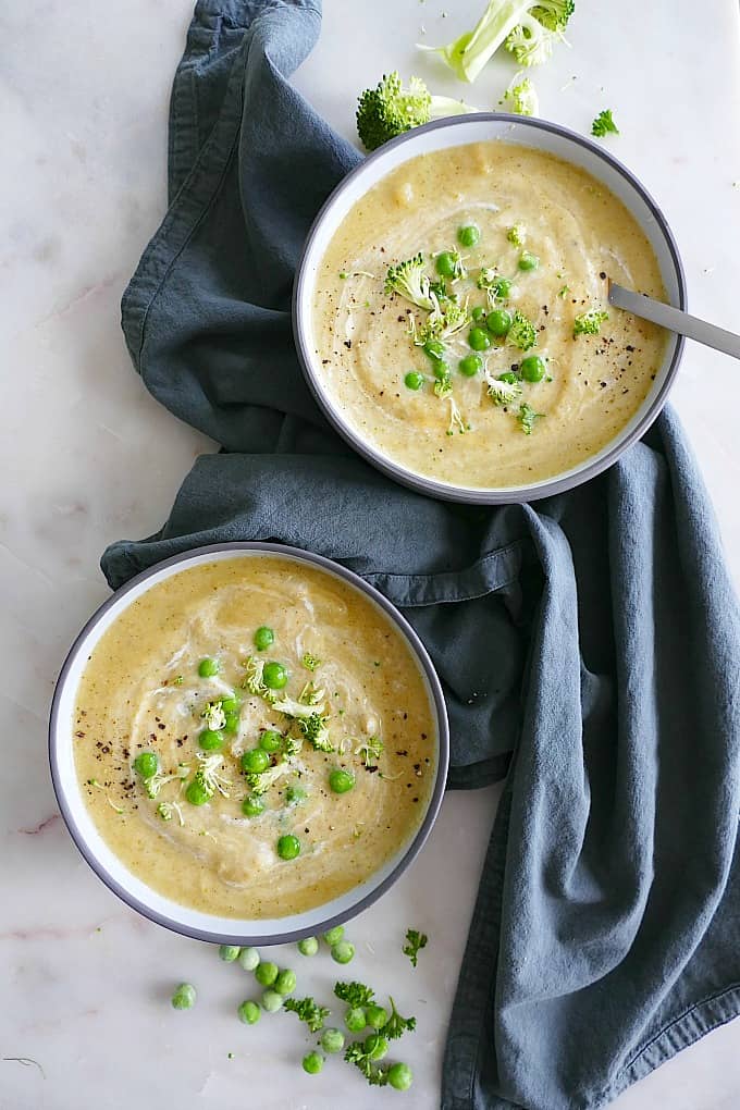 two bowls of broccoli and potato soup on a blue napkin on a white counter