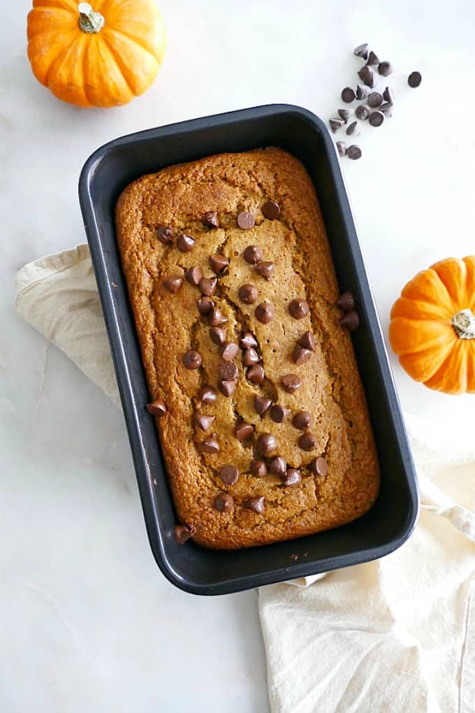 A loaf of banana pumpkin oatmeal bread in a black bread pan next to two mini pumpkins on a table. 
