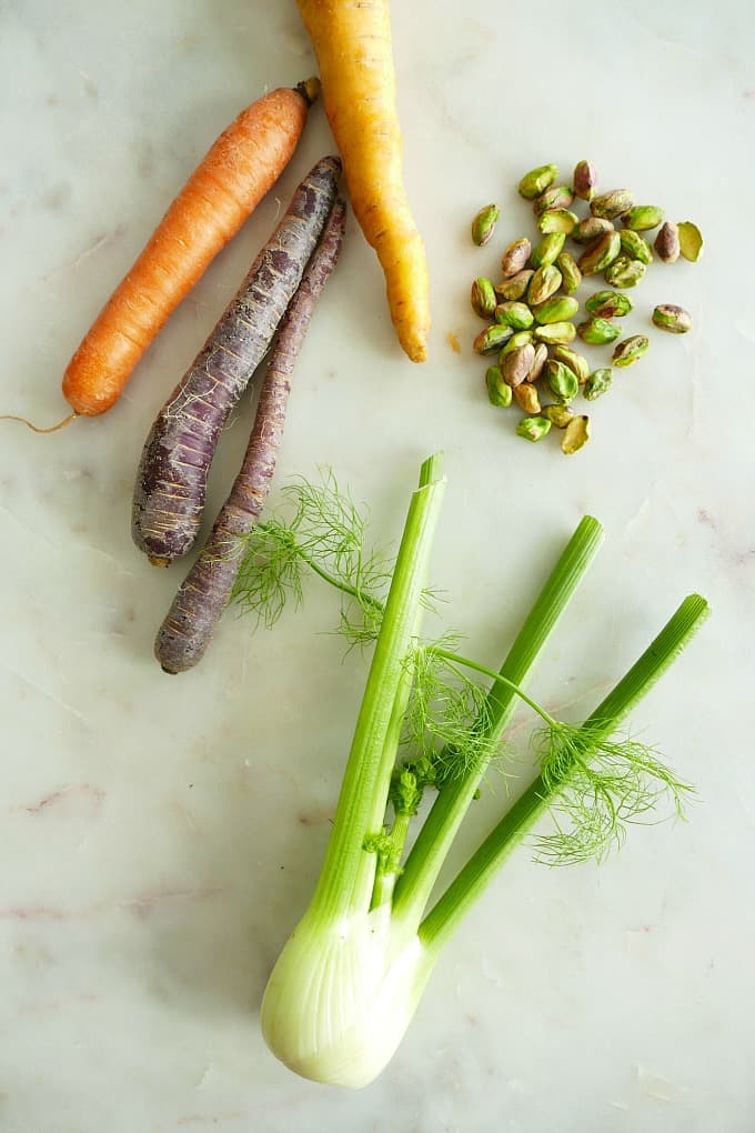 purple and orange carrots next to pistachios and a bulb of fennel on a counter