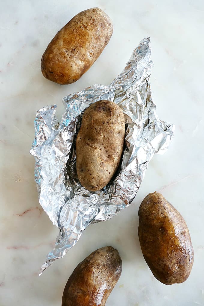 a baked potato on aluminum foil surrounded by 3 other potatoes on a counter