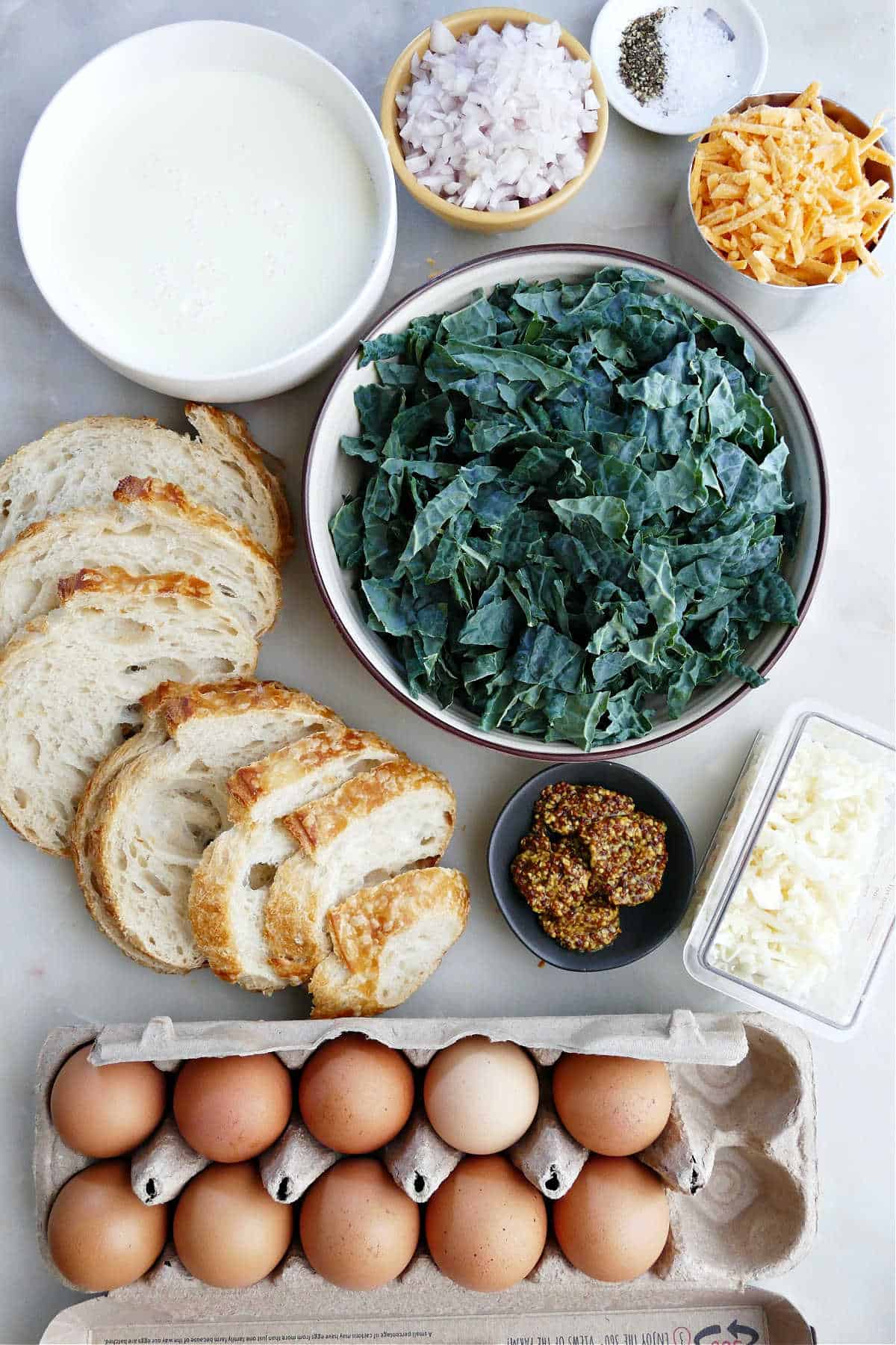 kale in a skillet next to bread cubes, cheese, and eggs on a counter