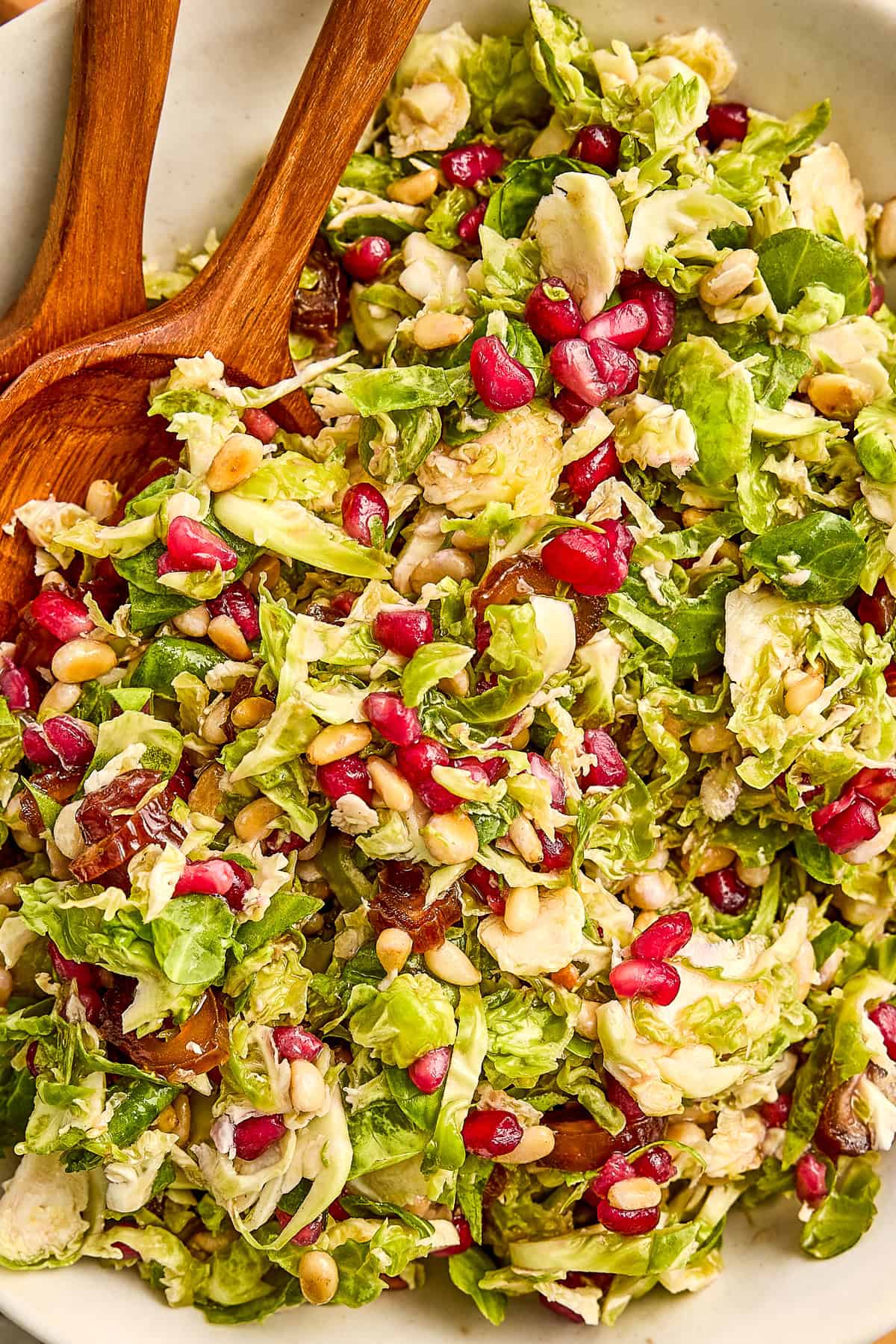 Shredded Brussels sprouts, pomegranates, and sliced dates in a salad bowl with tongs.