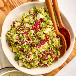 Brussels sprouts and date salad in a bowl with salad tongs on a wooden board.
