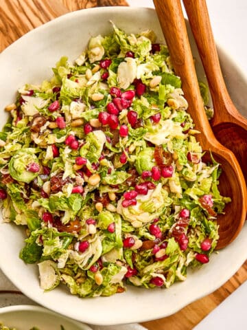 Brussels sprouts and date salad in a bowl with salad tongs on a wooden board.