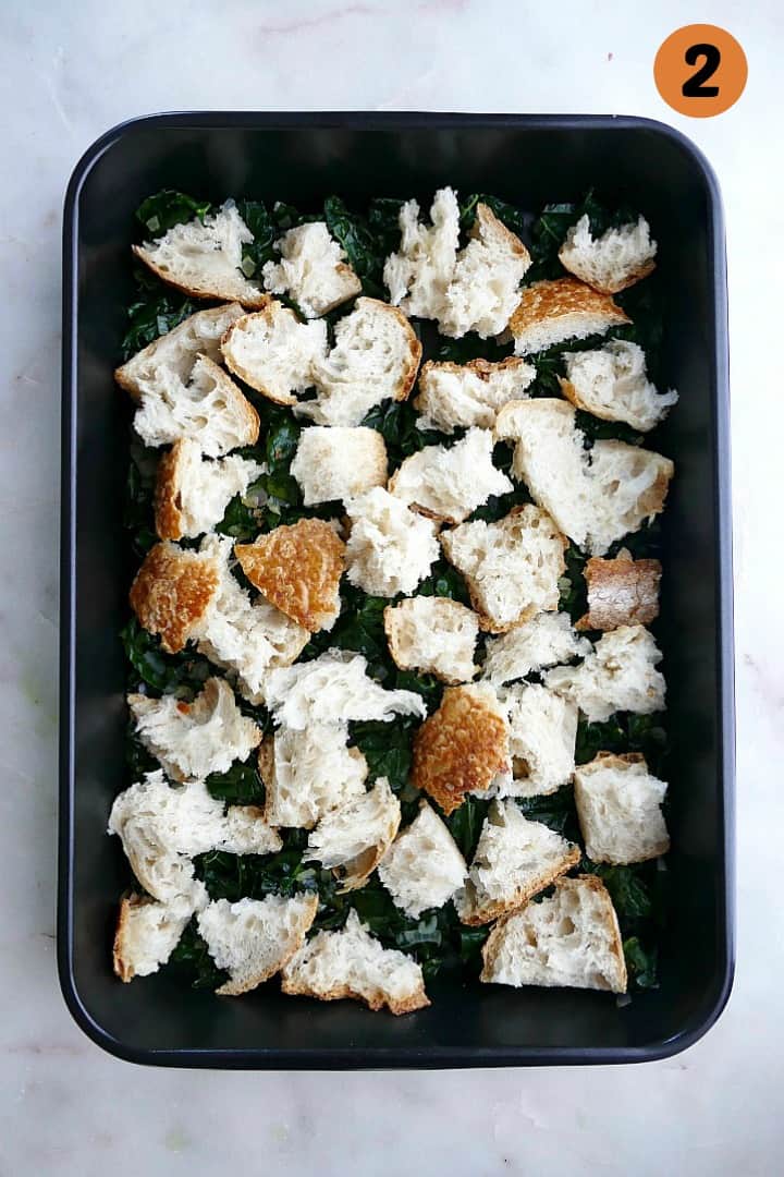 kale and shallots covered with sourdough bread cubes in a black 9x13 baking dish