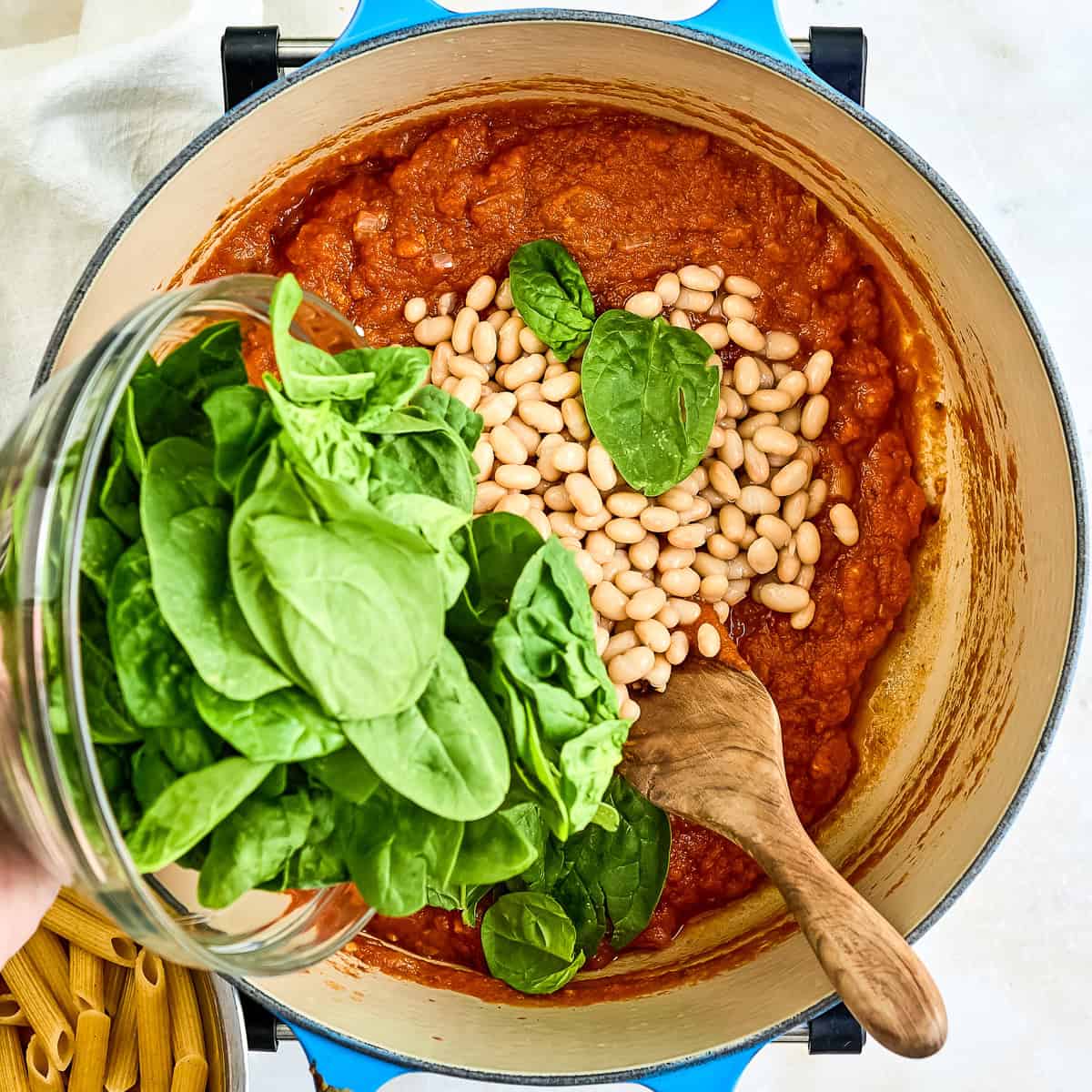 Woman adding baby spinach to a pot with pasta sauce and white beans.