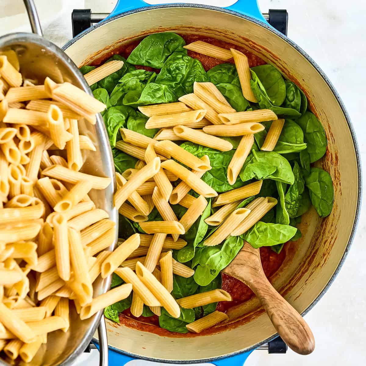Woman adding cooked pasta to a pot with sauce, spinach, and beans.