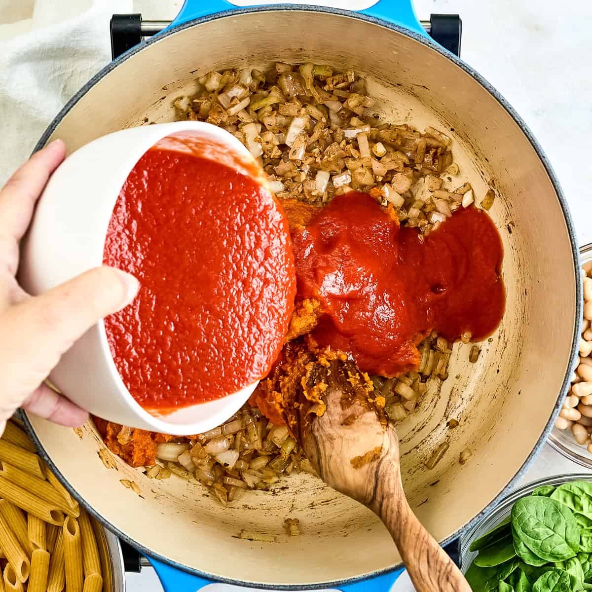 Woman adding tomato sauce to a pot with pumpkin, garlic, and onion.