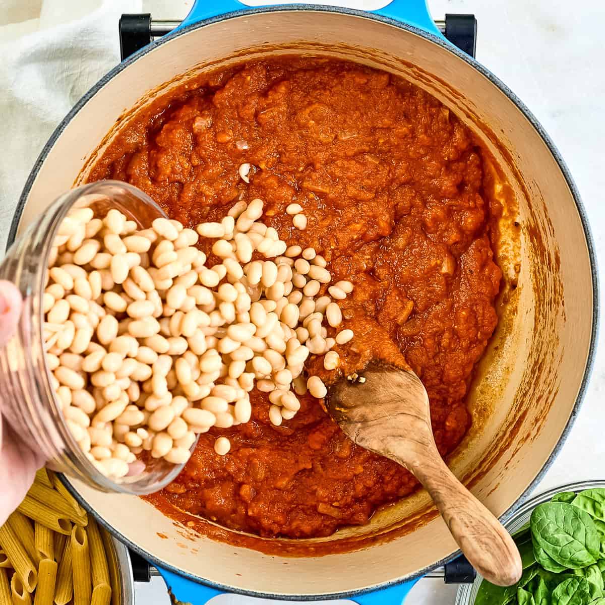 Woman adding drained white beans to a pot with pasta sauce.