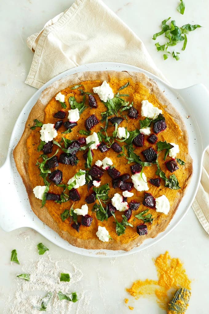 Butternut squash and beet pizza pizza on a round dish next to neutral kitchen towel.