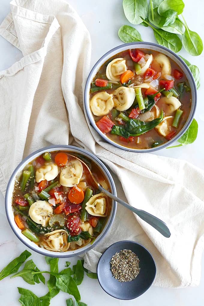two bowls of vegetable tortellini soup on a counter with a napkin and ingredients