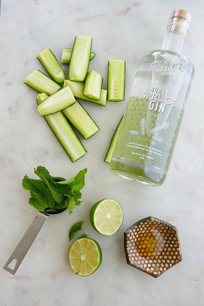 sliced cucumber, bottle of gin, mint leaves, lime, and honey spread out on a counter