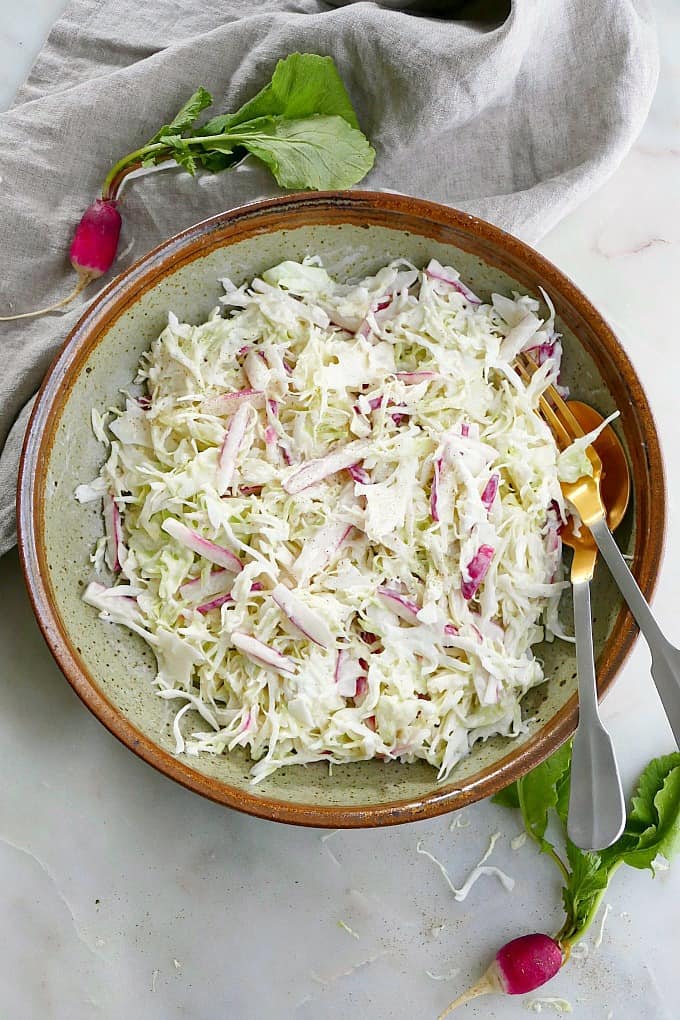 coleslaw and sliced radishes in a large bowl on a counter with a fork and spoon