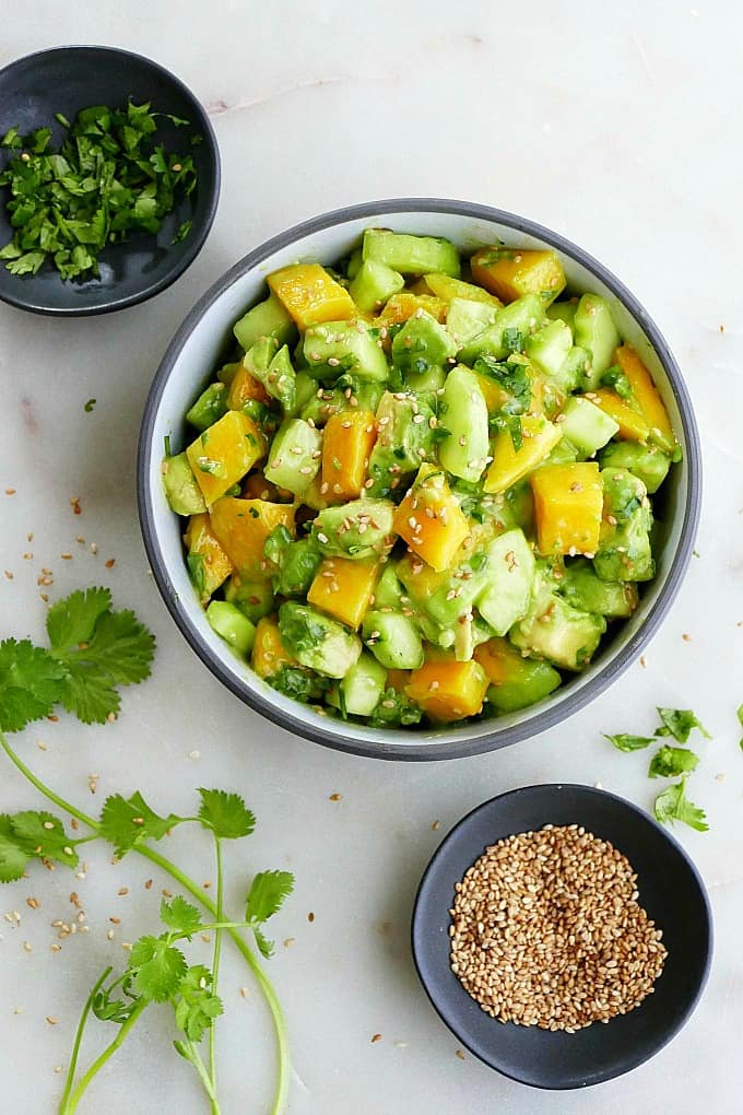 mango cucumber salad in a bowl on the counter next to sesame seeds and cilantro