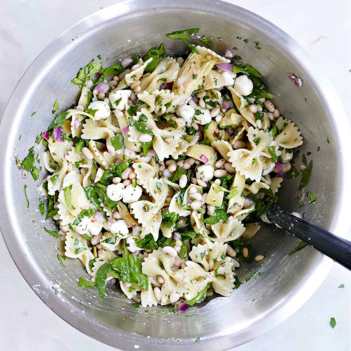 pasta salad with artichokes and spinach in a mixing bowl with a spoon on a counter