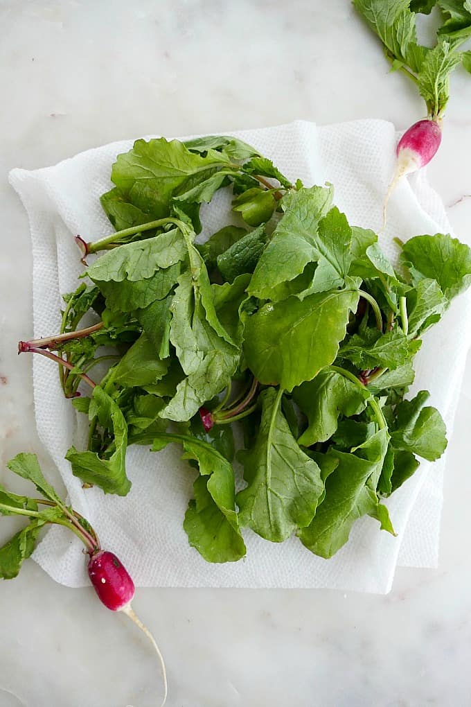 radish leaves on a damp paper towel on a counter next to full radishes