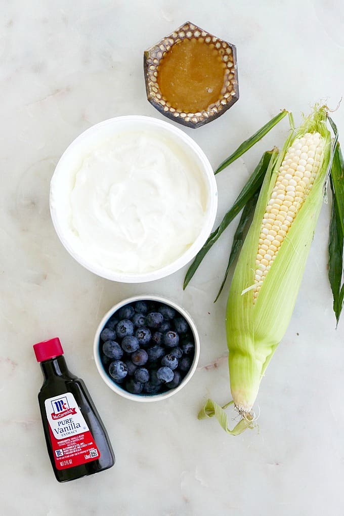 vanilla extract, yogurt, blueberries, sweet corn, and honey spread out on a counter