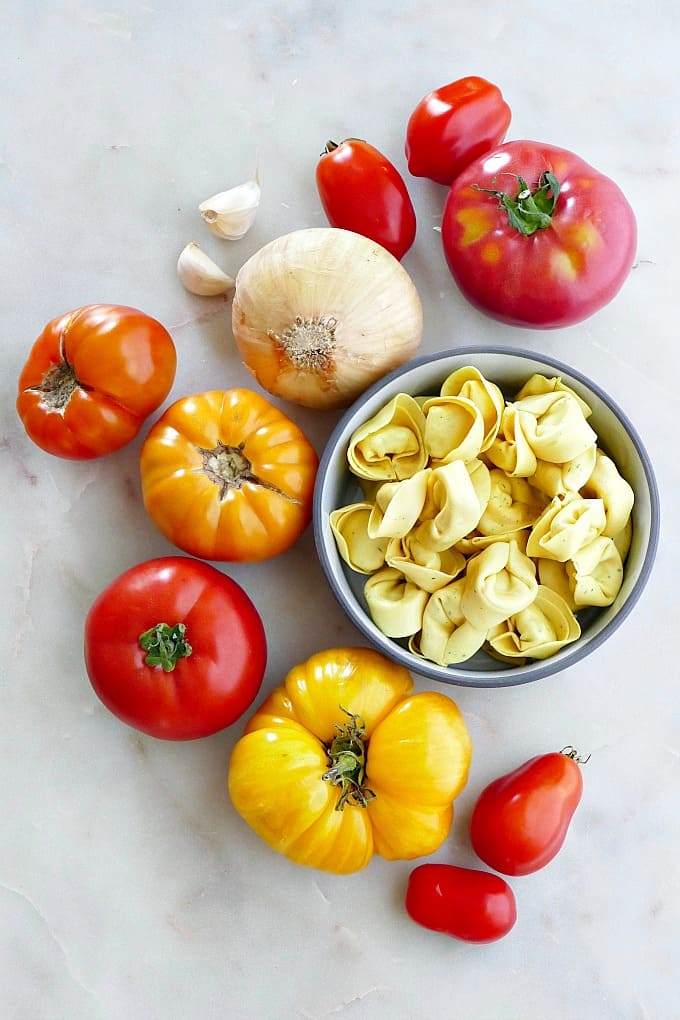 heirloom tomatoes, tortellini, sweet onion, and garlic cloves next to each other on a counter