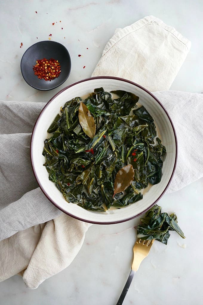 collard greens in white bowl with pepper flakes and fork on white counter