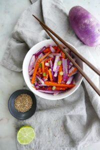 purple radish salad with carrots and green onions in a white bowl on top of a grey napkin with brown chopsticks on the top of the bowl