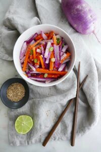 purple radish salad with carrots and green onions in a white bowl on top of a grey napkin next to brown chopsticks, sesame seeds, a purple radish, and a lime