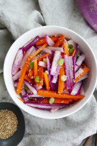 purple radish salad with carrots and green onions in a white bowl on top of a grey napkin