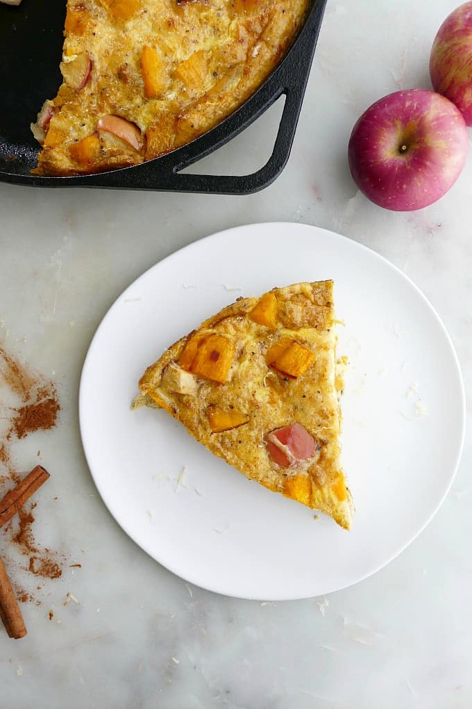 slice of apple pumpkin frittata on a white plate next to apples and a skillet