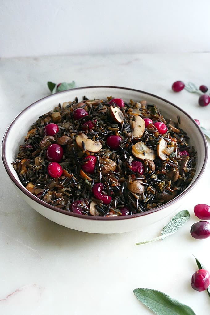 side view of wild rice pilaf in a white bowl with sage and cranberries on the side