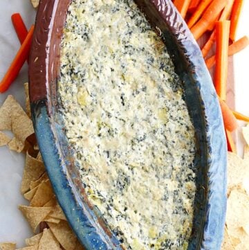 spinach artichoke dip with greek yogurt in a blue and brown dish surrounded by carrots and chips