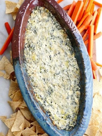 spinach artichoke dip with greek yogurt in a blue and brown dish surrounded by carrots and chips