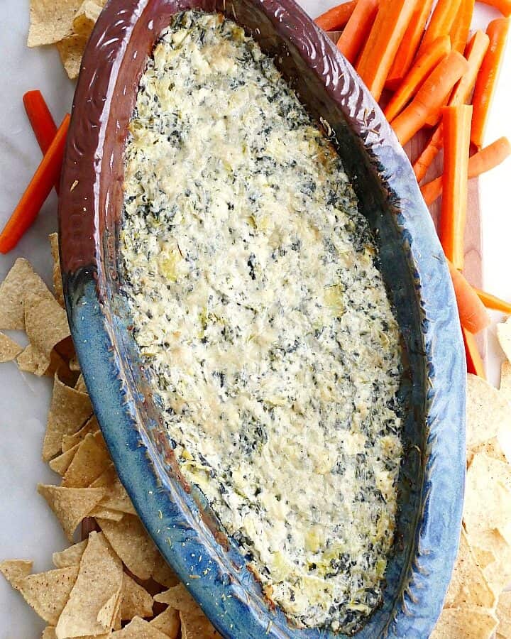 spinach artichoke dip with greek yogurt in a blue and brown dish surrounded by carrots and chips