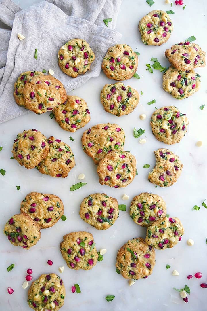 white chocolate chip spinach cookies on a white counter with spinach, pomegranates, and chips