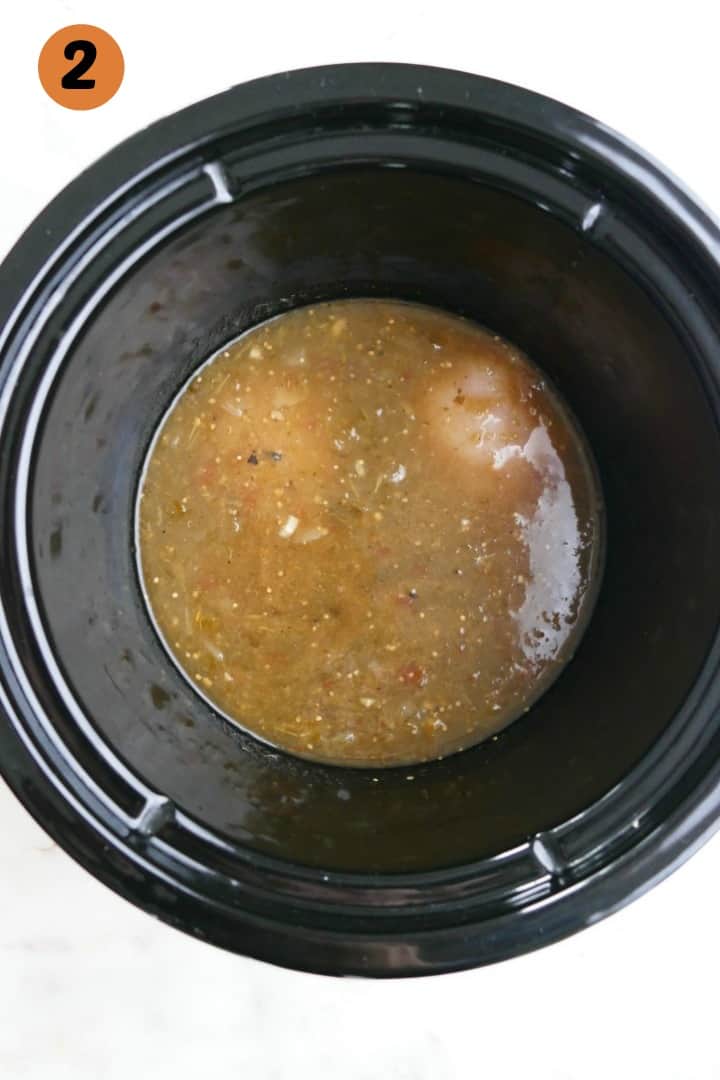 two chicken breasts covered with a jar of salsa verde in a crockpot on a white counter