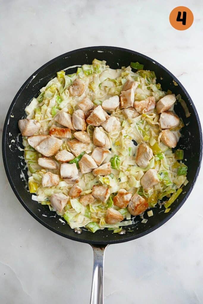sliced leeks, cream sauce, and diced chicken cooking in a black skillet on a white counter